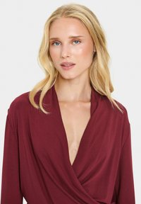 Burgundijas krāsas apņēmuma tops ar garām piedurknēm un dziļu V-veida kaklu. Mīksts, gluds audums ar pludinātām detaļām. Raksturo eleganta siluets.