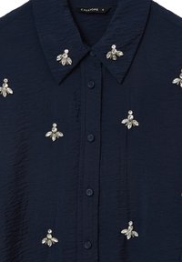 Camicia blu navy con tessuto testurizzato, caratterizzata da accenti decorativi in argento e numerosi abbellimenti simili a cristalli sul davanti e sul colletto.