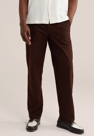 Mężczyzna ubrany w ciemnobrązowe spodnie, białą koszulę i czarno-białe skórzane mokasyny, stojący z jedną ręką w kieszeni na jednolitym tle.