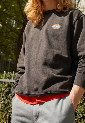 Osoba nosząca wyblakłą czarną bluzę Dickies na czerwonej koszuli i jasnoszarych spodniach, stojąca na zewnątrz z rękami w kieszeniach.