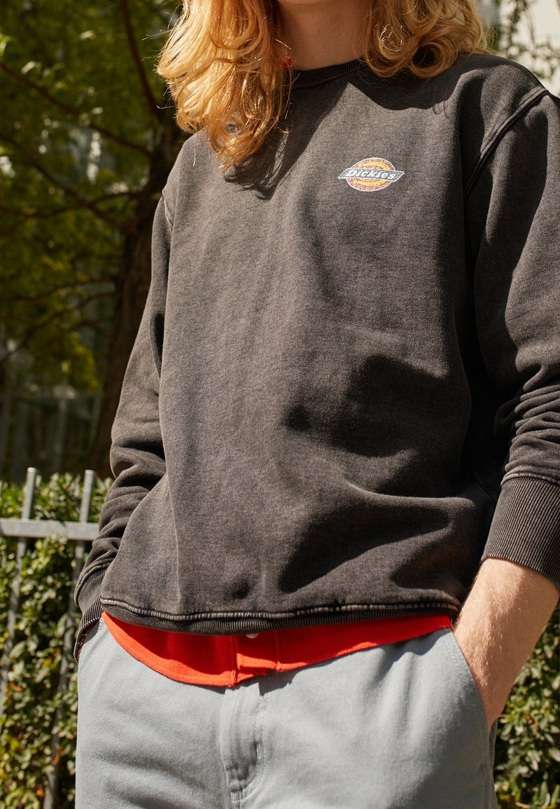 Personne portant un sweat-shirt noir délavé Dickies par-dessus une chemise rouge et un pantalon gris clair, debout à l'extérieur avec les mains dans les poches.