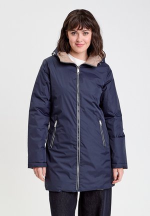 Femme portant une longue veste zippée bleu marine avec une doublure de capuche beige, se tenant devant un fond blanc uni.