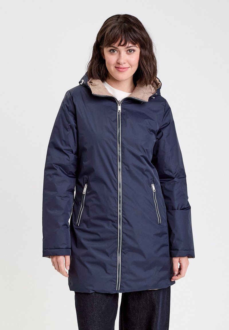 Femme portant une longue veste zippée bleu marine avec une doublure de capuche beige, se tenant devant un fond blanc uni.