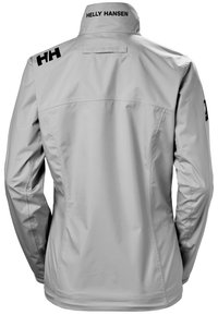 Chaqueta impermeable gris claro con un cuello alto, que presenta logotipos negros en la espalda y el hombro. Textura suave y diseño ajustado.