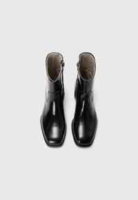 Botas de cuero negro hasta el tobillo con un acabado brillante, diseño de puntera cuadrada, cremallera lateral y detalles de costura mínimos. El interior es beige.