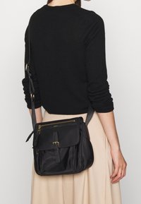 Žena v černém topu s dlouhým rukávem a béžové sukni nese černou crossbody tašku s několika zipovými oddíly a předním sponou.