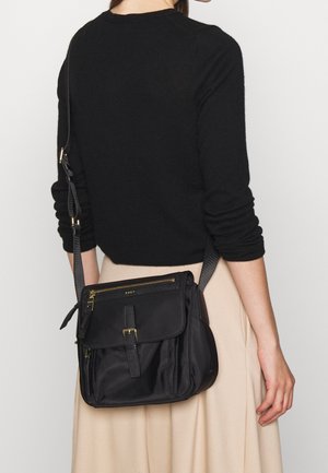 Una donna con un top nero a maniche lunghe e una gonna beige porta una borsa a tracolla nera con più scomparti con zip e una fibbia frontale.