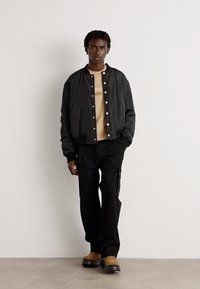 Black bomber jacket with snap buttons, beige crew neck shirt underneath, black pants, and brown footwear. Casual, relaxed fit.