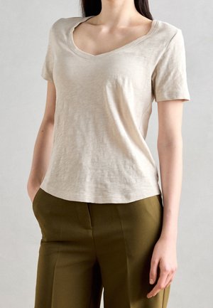 Kobieta ubrana w beżową koszulkę z krótkim rękawem i dekoltem w serek oraz oliwkowe spodnie, stojąca z jedną ręką w kieszeni na jednolitym tle.