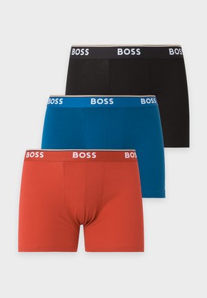 Uomo magro con pelle scura, in piedi a piedi nudi, che indossa boxer blu con elastico bianco con marchio "BOSS", guarda leggermente di lato.