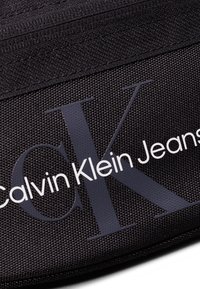 Juoda audinio rankinė su tekstūruotu paviršiumi, dideliu pilku "CK" logotipu ir balta užrašu "Calvin Klein Jeans". Uždarymas užtrauktuku viršuje.