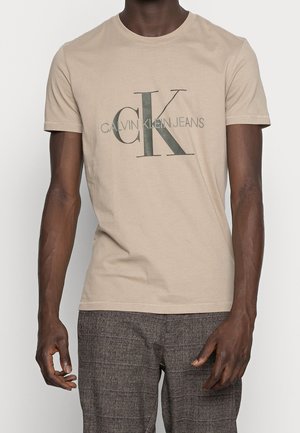 Osoba nosząca beżowy t-shirt z krótkim rękawem z dużym tekstem "CK" i "Calvin Klein Jeans", w zestawieniu z szarymi spodniami w kratę.