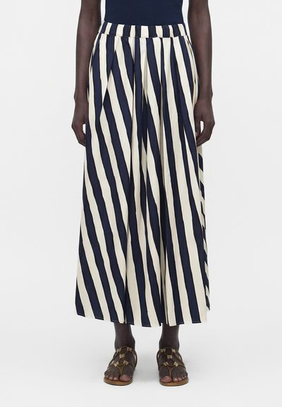 Personne à la peau noire portant un pantalon large avec des rayures diagonales crème et bleu marine et des sandales marron, debout devant un fond blanc.