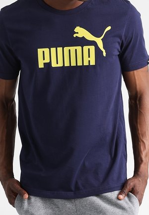 Dunkelblaues T-Shirt mit gelbem Puma-Logo und Schriftzug, getragen von einer Person mit den Händen in den Taschen einer grauen Hose.