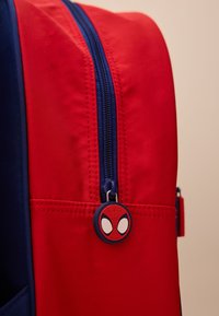 Red and blue backpack with a smooth texture, featuring a zipper and a Spider-Man logo pull tab. Compact design for easy carrying.