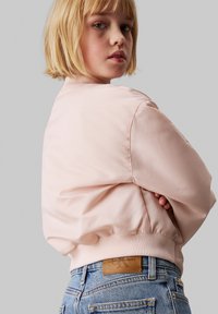 Chaqueta bomber de color rosa claro con puños y dobladillo acanalados, textura suave, llevada sobre unos vaqueros de cintura alta azules que cuentan con un parche de cuero de la marca.