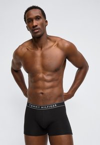 Μυώδης άνδρας που στέκεται με τα χέρια πίσω από την πλάτη, φορώντας μαύρα μπόξερ Tommy Hilfiger σε απλό ανοιχτό γκρι φόντο.