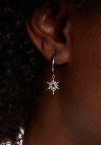 Pendiente en forma de estrella plateada con una piedra negra central, colgando de un aro. El diseño presenta acentos puntiagudos y un acabado pulido.