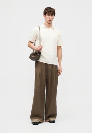 Nuori mies, jolla on teksturoitu valkoinen lyhythihainen paita, leveät ruskeat housut, beiget kengät ja ruskea nahkainen olkalaukku.