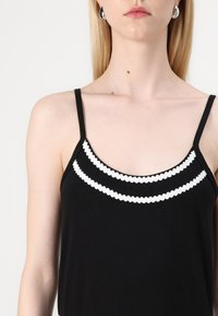 Sort camisole med tynde stropper og dekorativ hvid bølget kant langs udskæringen. Glat stoftekstur.