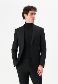 Black tailored suit with a slim fit, featuring a single-breasted jacket and matching trousers, worn over a black turtleneck. Smooth texture.