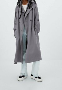 Grauer Trenchcoat mit doppelter Knopfleiste, großen Revers und Seitentaschen. Getragen über hellblauen Jeans und schwarzen Sneakers.