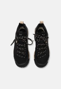 Ein Paar schwarze Sportschuhe mit beigen Akzenten, gemusterten Schnürsenkeln, die zu Schleifen gebunden sind, und Zuglaschen auf weißem Hintergrund.