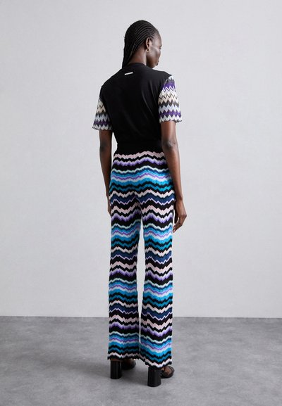 Μοντέρνο σύνολο που περιλαμβάνει ένα μαύρο τοπ με μοτίβο κοντά μανίκια, συνδυασμένο με φαρδιά παντελόνια με ζιγκ-ζαγκ σχέδιο σε μπλε, μωβ και μαύρο.