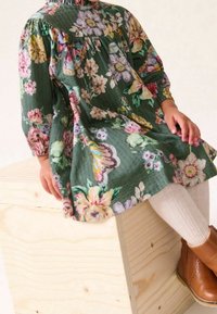 Blumenkleid aus grünem Stoff mit langen Ärmeln, einem gerafften Ausschnitt und bunten Blumenmustern. Braune Lederstiefeletten und helle Strumpfhosen.