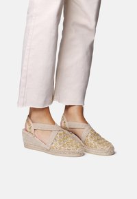 Espadrilles de cuña en color beige con un patrón tejido dorado, con tiras entrecruzadas y una suela de yute texturizada. Adecuadas para uso casual.