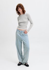 Light gray long-sleeve shirt with white trim, paired with loose light blue jeans. Model stands with one hand in pocket, wearing black slip-on shoes.