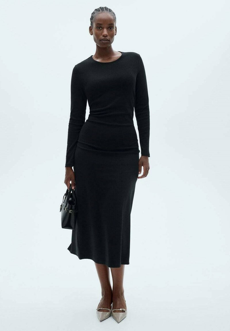 Black fitted long-sleeve dress with a round neckline, smooth texture, and a mid-length skirt. Accessories include a small black handbag.