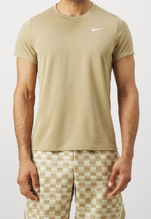 Man wearing a tan Nike short-sleeve shirt and beige checkered shorts, standing against a plain white background.