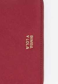 Cartera de tela roja con el logotipo dorado en relieve "BIMBA Y LOLA", textura suave y costura amarilla en contraste a lo largo de la cremallera.