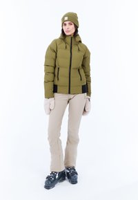 Olivgrüner Pufferjacke mit Reißverschluss, elastischen Bündchen und Seitentaschen; kombiniert mit beigen Skihosen und schwarzen Skistiefeln. Beigefarbene Fäustlinge inklusive.