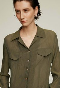 Blusa ligera color verde oliva, abotonada y con cuello, dos bolsillos en el pecho y una tela suave y texturizada. Brillo sutil y corte relajado.