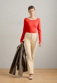 Piros hosszú ujjú felső, krém színű bőrszárú nadrág, sötétszürke bőrkabátot tart. Világos színű cipők egészítik ki az outfitet. Minimalista háttér.