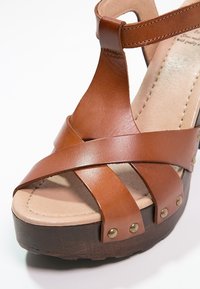 Sandale plateforme en cuir marron avec des brides croisées, une semelle en bois robuste et des clous métalliques le long du bord pour plus de détails.