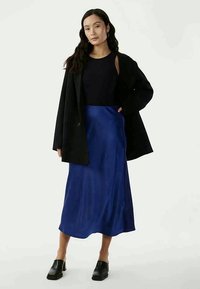 Musta ylipitkä takki, tummansininen satiininen liukuhame, slim fit musta hihaton paita ja mustat puukengät. Sileät pinnat rentouttavalla siluetilla.