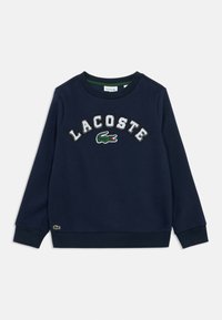 Tumši zils Lacoste sporta krekls ar ribotām aprocēm, apļa kakla izgriezumu, balti izšūtu zīmola nosaukumu un zaļu krokodilu logo uz krūšu un apakšmala.