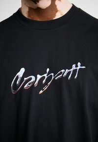 Zwarte katoenen t-shirt met een reflecterend, golvend logo-design in zilver, gecentreerd op de borst. Rond de hals en korte mouwen.