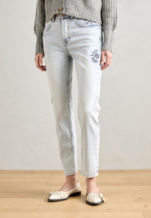 Jean délavé clair avec petite broderie soleil sur la cuisse, porté avec un cardigan en maille gris et des chaussures plates beige avec détail boucle.