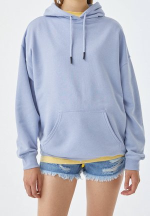 Personne portant un sweat à capuche oversize violet clair avec poche avant, superposé à un t-shirt jaune, associé à un short en jean déchiré.