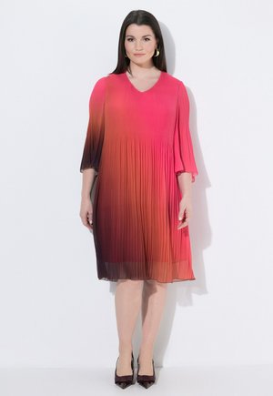 Kvinde stående mod hvid baggrund iført en plisseret, ombre pink til mørk lilla knælængde kjole og sorte spidse hæle.