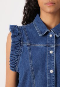 Denimskjorte med krage, perleknapper og rynkede skulderdetaljer. Dyp blå farge med teksturert stofffinish.