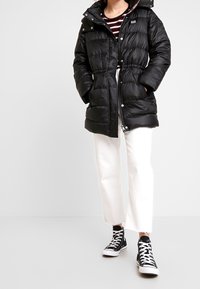 Chaqueta de plumas negra con cierre de botones y cremallera, diseño acolchado, cintura ajustable y capucha, llevada sobre pantalones cortos blancos y zapatillas negras.