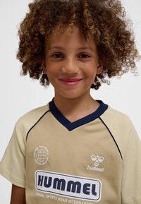 Uśmiechnięte dziecko z kręconymi włosami, ubrane w beżową sportową koszulkę z granatowymi lamówkami i logo marki "Hummel" na jednolitym białym tle.