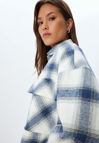 Camisa de cuadros azul y blanca con una tela gruesa, que presenta un cuello, un bolsillo frontal y un patrón texturizado, con un corte holgado.