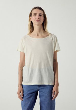 Pitkähiuksinen vaaleatukkainen nainen, joka pukeutuu yksinkertaiseen off-white -väriseen lyhythihaiseen paitaan ja sinisiin farkkuihin, seisoo yksiväristä vaaleanharmaata taustaa vasten.