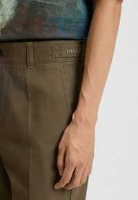 Rechterhand rustend naast de persoon in olijfgroene Hugo-broek en een kleurrijk shirt tegen een eenvoudige achtergrond.